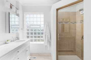 A bathroom with a white cabinet and countertop, glass block window, and a shower with brown tiles.