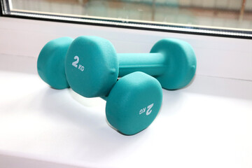 Two 2-kilogram sea-green dumbbells lying on top of each other on a windowsill, close-up. Sports equipment for arm training. Sports, healthy lifestyle - horizontal color photo