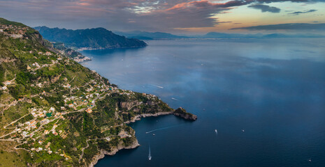 Fototapeta premium Aerial view of the Amalfi Coast in Italy with cliffs, green hills, terraced buildings, and boats on the Tyrrhenian Sea during sunset or sunrise.