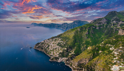 Aerial view of Amalfi, Italy, with cliffs, green hills, whitewashed buildings, a hillside hotel, and a vibrant sunset over the Tyrrhenian Sea. © Aerial Film Studio