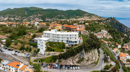 Obraz premium Aerial view of Amalfi, Italy, showing a white hotel on a rocky hill, lush greenery, terraced gardens, orange roofed houses, and the Tyrrhenian Sea.