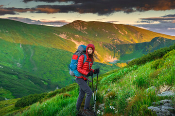 Fototapeta premium Female hiker in red jacket and hood standing on windy mountain slope at golden hour. Thoughtful gaze, trekking poles, dramatic light over green Carpathian hills in background