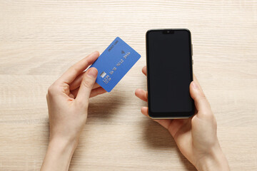 Woman with credit card and smartphone paying online at wooden table, top view