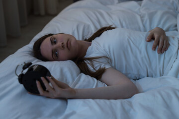 Frustrated young woman with alarm clock suffering from insomnia on bed indoors