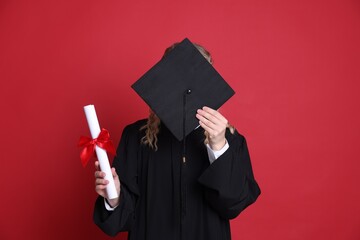 Student with diploma hiding behind mortarboard after graduation on red background