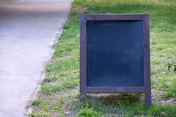 A blank chalkboard sign stands on lush green grass near a young tree in a sunny park. The landscape suggests an invitation for announcements, messages, or creative outdoor presentations.