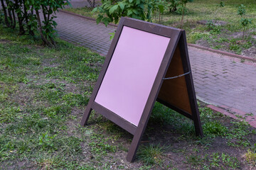 A blank pink chalkboard standing on a sidewalk outdoors, ready for business messages. The board is surrounded by greenery, making it ideal for attracting the attention of pedestrians passing by.