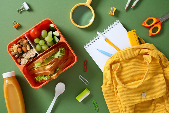 Healthy packed lunch for students: bento box with sandwich, grapes, and snacks, with school supplies and backpack.