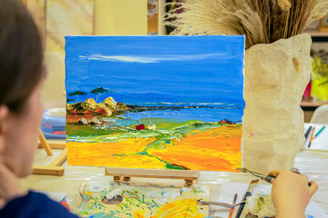 Young woman artist sitting near easel, painting picture with scenic sea landscape. Artist creating...