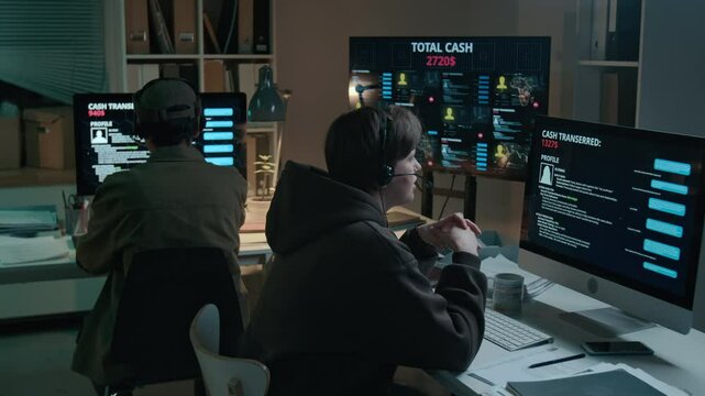 Medium full rear shot of young Caucasian female scam call center employee sitting in front of computer with victim profile, talking on phone while watching them transfer funds to fake safe account