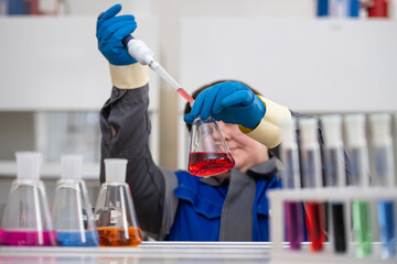 A female lab technician in a factory uniform and blue gloves pours red liquid from a pipette into a flask. Blurred laboratory flasks with colorful liquids emphasize the vibrant analysis process.