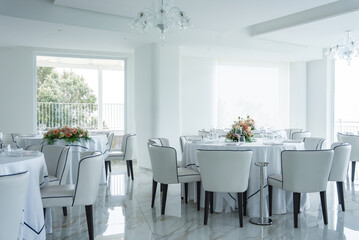 Modern dining space with white walls, marble floors, round tables, floral centerpieces, and large windows offering partial views of greenery and sea.