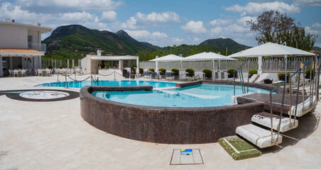A modern outdoor pool area with a circular pool, white umbrellas, lounge chairs, and potted plants, set against green hills and a partly cloudy sky.