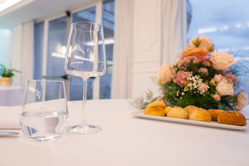 A white table features a glass of water, a wine glass, bread rolls, and a floral arrangement. Large glass doors reveal an evening sky in Amalfi, Italy.