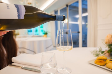 A bottle of white wine is poured into a glass on a table with white linens, pastries, and a floral centerpiece in a bright Amalfi restaurant interior.