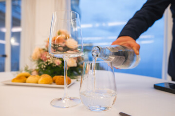 A hand pours water into a glass on a refined table with bread rolls, floral centerpiece, and wine glass, set against a sea and sky view in Amalfi.