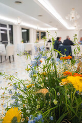 Bright dining space with white decor, modern chandeliers, vibrant floral centerpiece, polished floors, and people seated at tables in conversation.