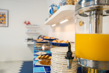 A breakfast buffet in a luxury Amalfi hotel features orange juice, pastries on blue dishes, white plates, and decorative pottery with red coral accents.