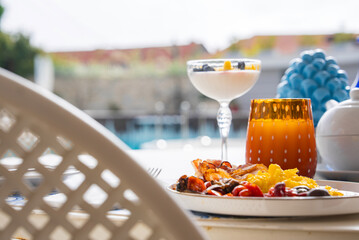 A vibrant breakfast table with eggs, vegetables, juice, and yogurt. Blurred pool area, stone wall, greenery, and red roofed buildings in the background.