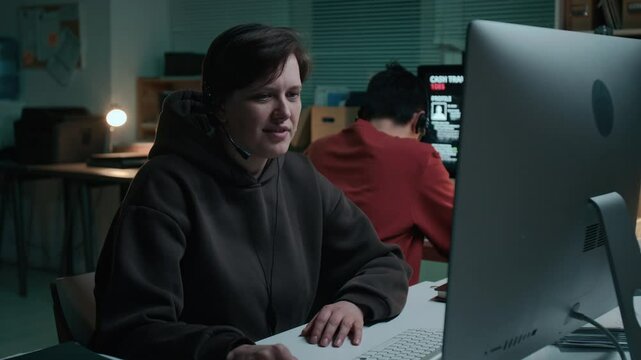 Medium shot of young Caucasian female fraudster talking politely to unsuspecting victim at scam call center, signing them up for fake investment scheme, hanging up, cheering and drumming on desk