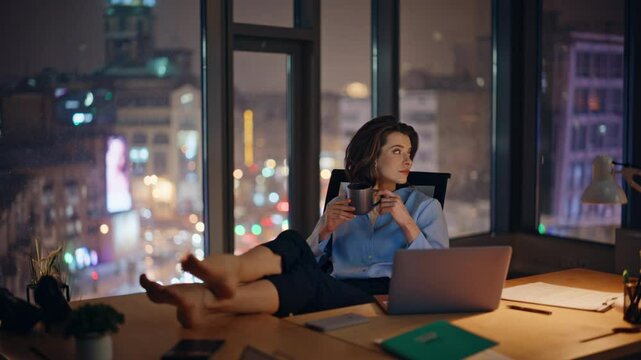 Relaxed overtime worker resting in night office putting bare feet on work desk. 