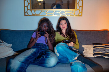 Two young women are relaxing on a comfortable sofa, enjoying a videogame session together in their...