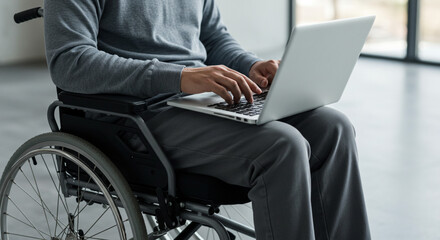 Man in wheelchair using laptop, wearing grey sweater and pants, indoors near window, working remotely online