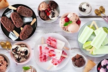 Cool summer food table scene. Assortment of ice cream bowls, popsicles and frozen treats. Overhead view on a white wood background.