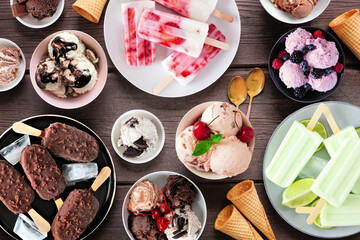 Cool summer food table scene. Assortment of ice cream bowls, popsicles and frozen treats. Above view on a dark wood background.