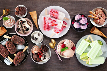 Cool summer food table scene. Variety of ice cream bowls, popsicles and frozen treats. Top view on a dark wood background.