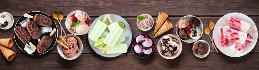 Cool summer food table scene. Assortment of ice cream bowls, popsicles and frozen treats. Top down view on a dark wood banner background.