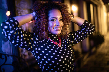 Black woman with curly hair in a relaxed pose with hands on her head, in a neon-lit night scene...