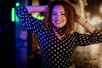 Fototapeta premium Black woman with curly hair smiling confidently in a neon-lit street scene inspired by the 1980s. Her striking look with a polka-dot blouse and sparkly skirt conveys style, joy, and bold attitude.