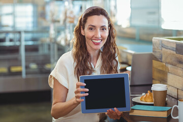 Woman sitting at counter in café holding tablet with blank screen beside croissant and coffee cup