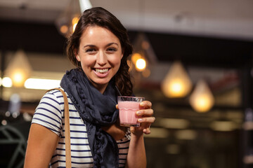Woman standing in cafe under lamps holding cup of pink drink with straw and tan-strap bag