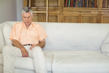 Senior man wearing orange-striped shirt sitting on sofa in living room near bookcase, copy space