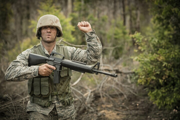 Fototapeta premium Soldier wearing camo helmet vest standing in forest clearing raising arm gripping assault rifle