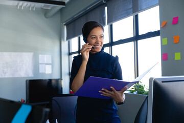 Late-twenties woman speaking on wireless headset and holding folder in office with monitors, prints