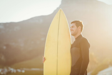 Male surfer wearing black wetsuit standing on beach holding yellow surfboard during golden hour