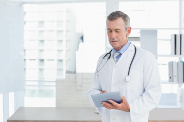Mature male doctor wearing lab coat reviewing tablet in clinic with stethoscope at desk, copy space