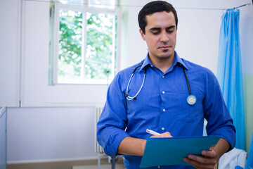 Doctor writing notes on blue clipboard wearing stethoscope in clinic by window foliage and curtain