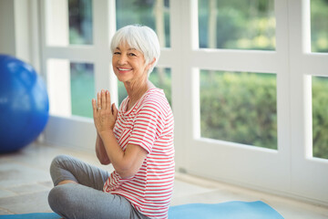 Senior woman wearing workout clothes sitting on blue mat beside exercise ball at home near windows