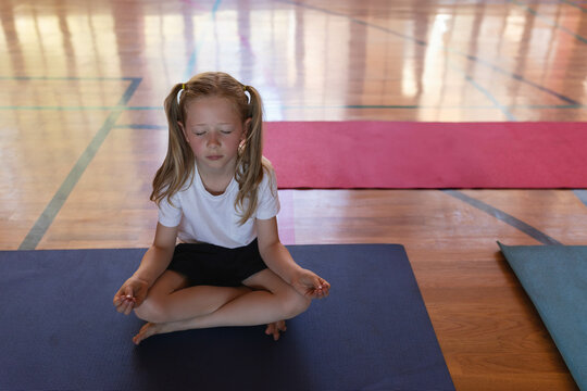 Fototapeta Girl sitting cross-legged on blue yoga mat on polished wooden hall floor, near red, green mats
