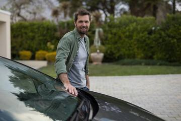 Mid adult man standing beside dark car on paved driveway wearing olive green shirt near planter