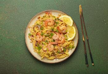 Pad thai noodles garnished with shrimp, peanuts, green onions, lemon wedges, and served with chopsticks on a rustic green background, Thai cuisine
