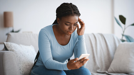 Worried black lady looking at smartphone screen, sitting on couch at home, concerned african american woman waiting for important call, reading unpleasant sms, spending time with phone in living room