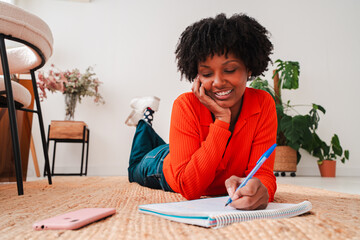 A cheerful young woman is lying on the floor and writing in her notebook, reflecting her creativity and enthusiasm for learning as she engages in productive writing and thought exploration at home.