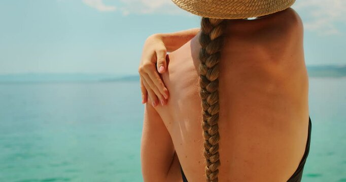 Woman in swimsuit and sunhat applies sunscreen to her shoulder on a sunny day