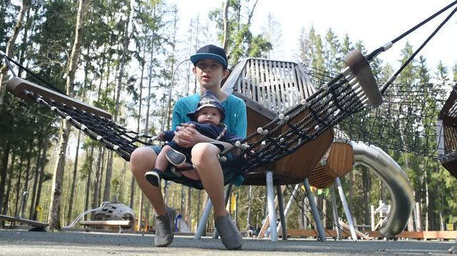 A 12 year old boy with his baby brother on his knees swings on a hammock swing at a playground in Borovsky Park in Moscow.	