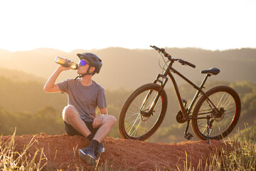 Young man resting and drinking water after cycling in nature at sunset. Concept of hydration,...